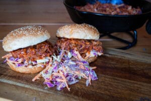 Twee pulled pork burgers met sesambroodjes en paarse coleslaw op houten snijplank met gietijzeren pan op achtergrond